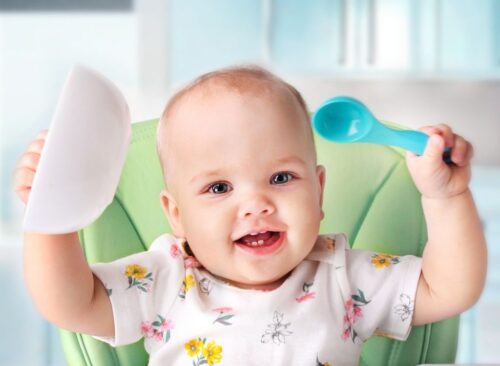 Baby spielt mit Löffel und Breischälchen und lacht in die Kamera
